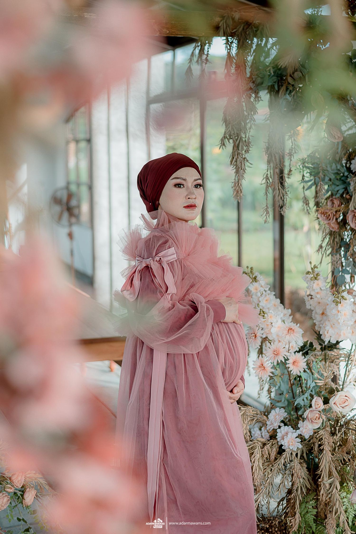 Expectant mother in pink ruffled dress and burgundy headpiece poses amid floral decorations.