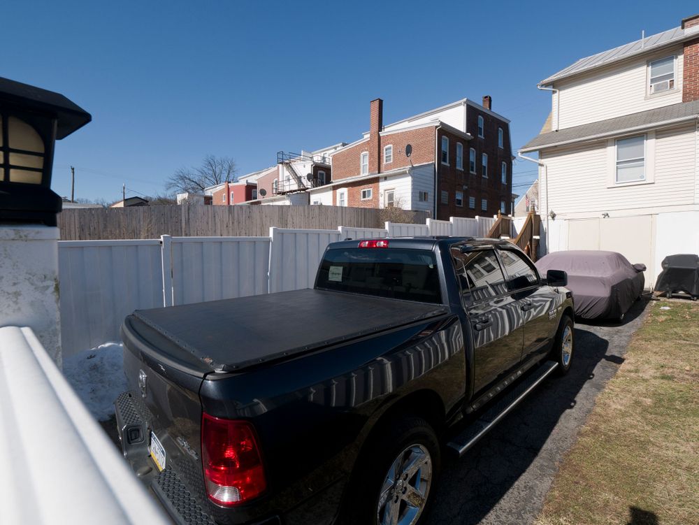 Black pickup truck parked in driveway next to white fence with urban buildings in background.
