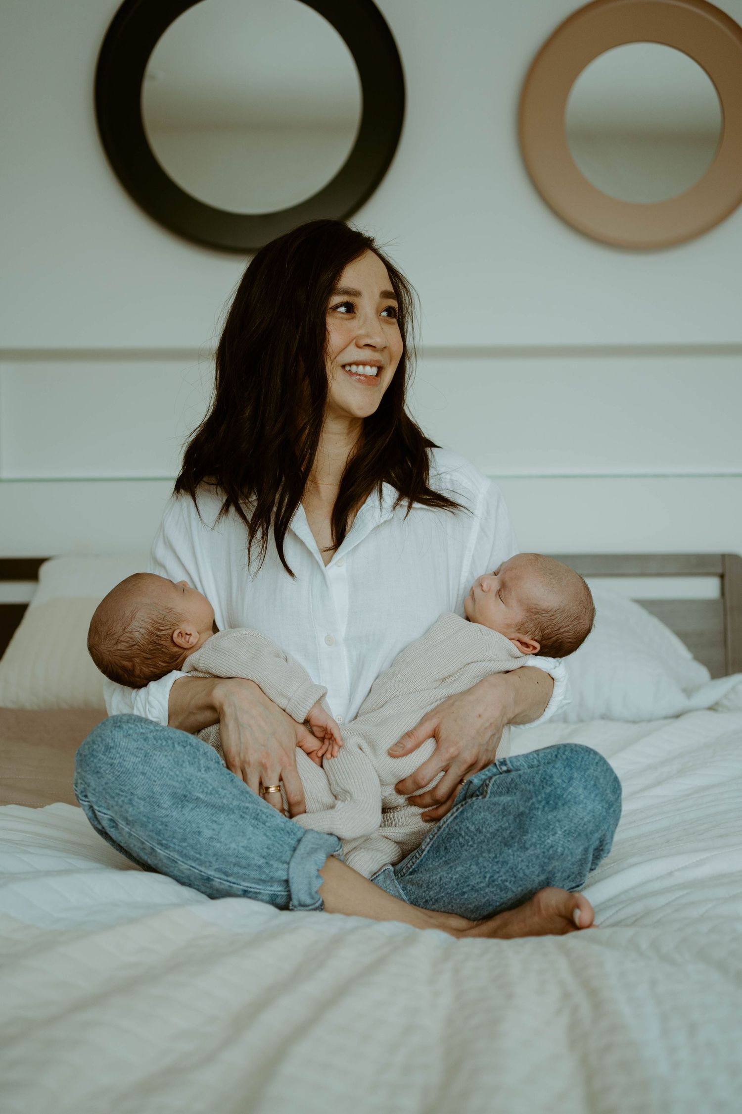 Mother on bed holding twin babies while sitting cross-legged in white button-down shirt.
