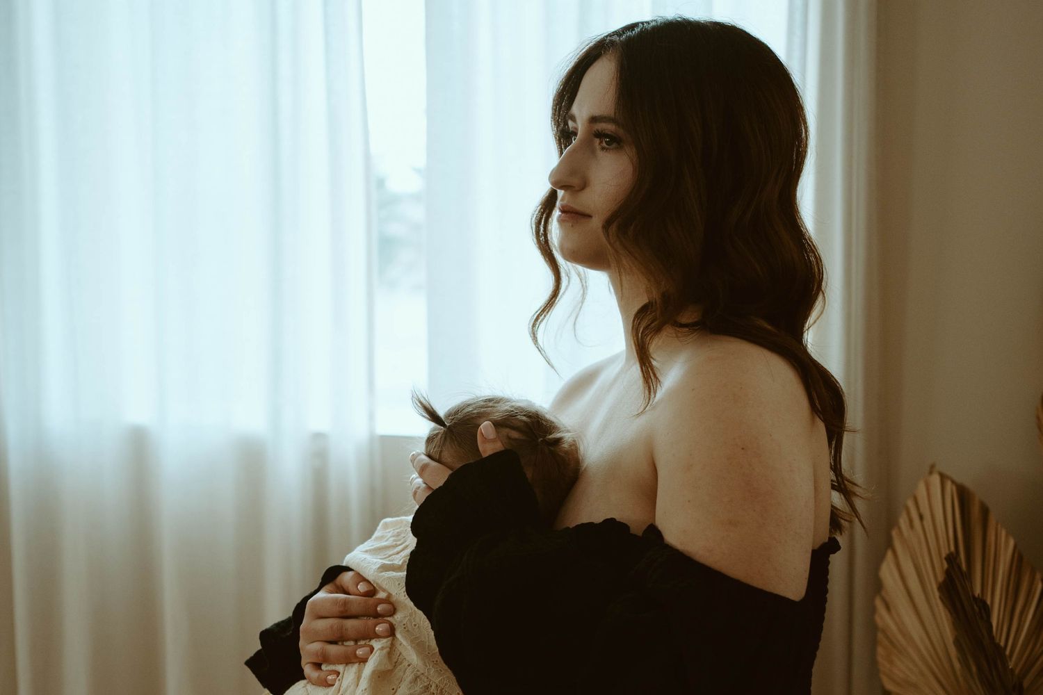 Soft natural light streams through window onto mother cradling newborn in black outfit.