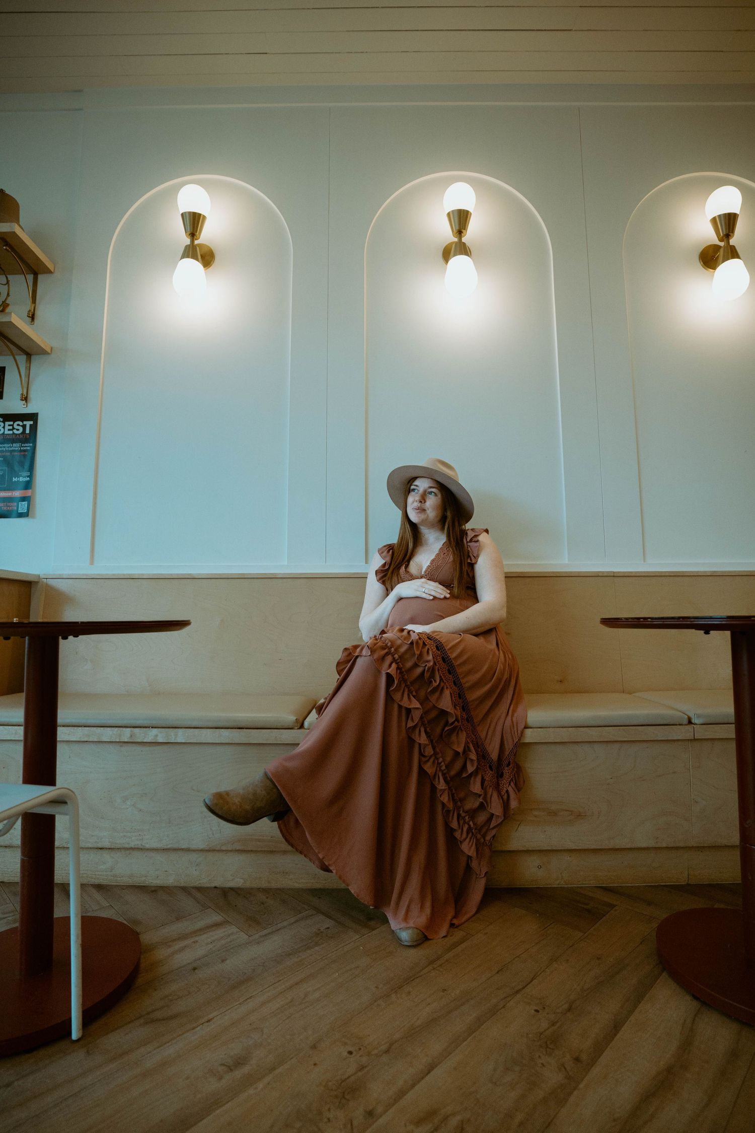Woman in brown dress and hat sits elegantly under arched wall sconces in rustic setting.