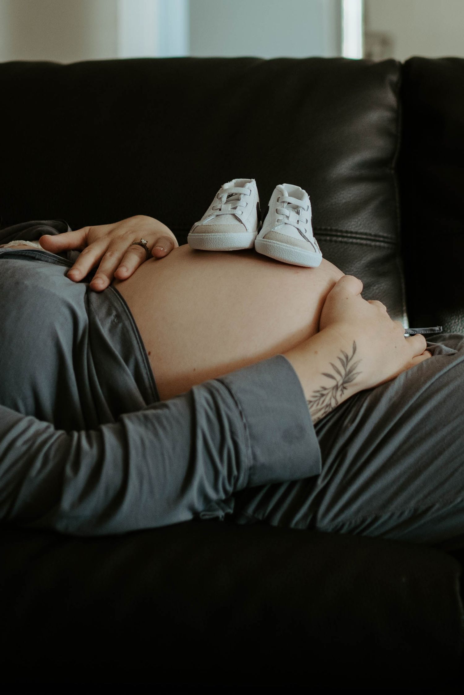 Small white baby shoes resting on pregnant belly on dark leather couch.