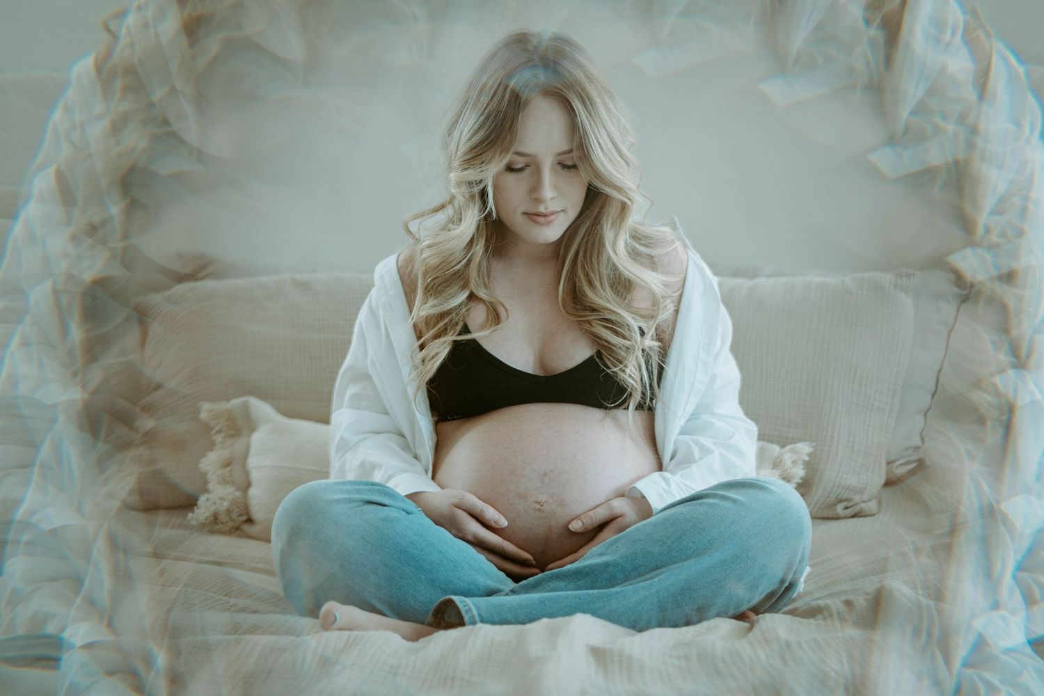 A maternity photo featuring a woman in a white cardigan and jeans sitting cross-legged on a couch.