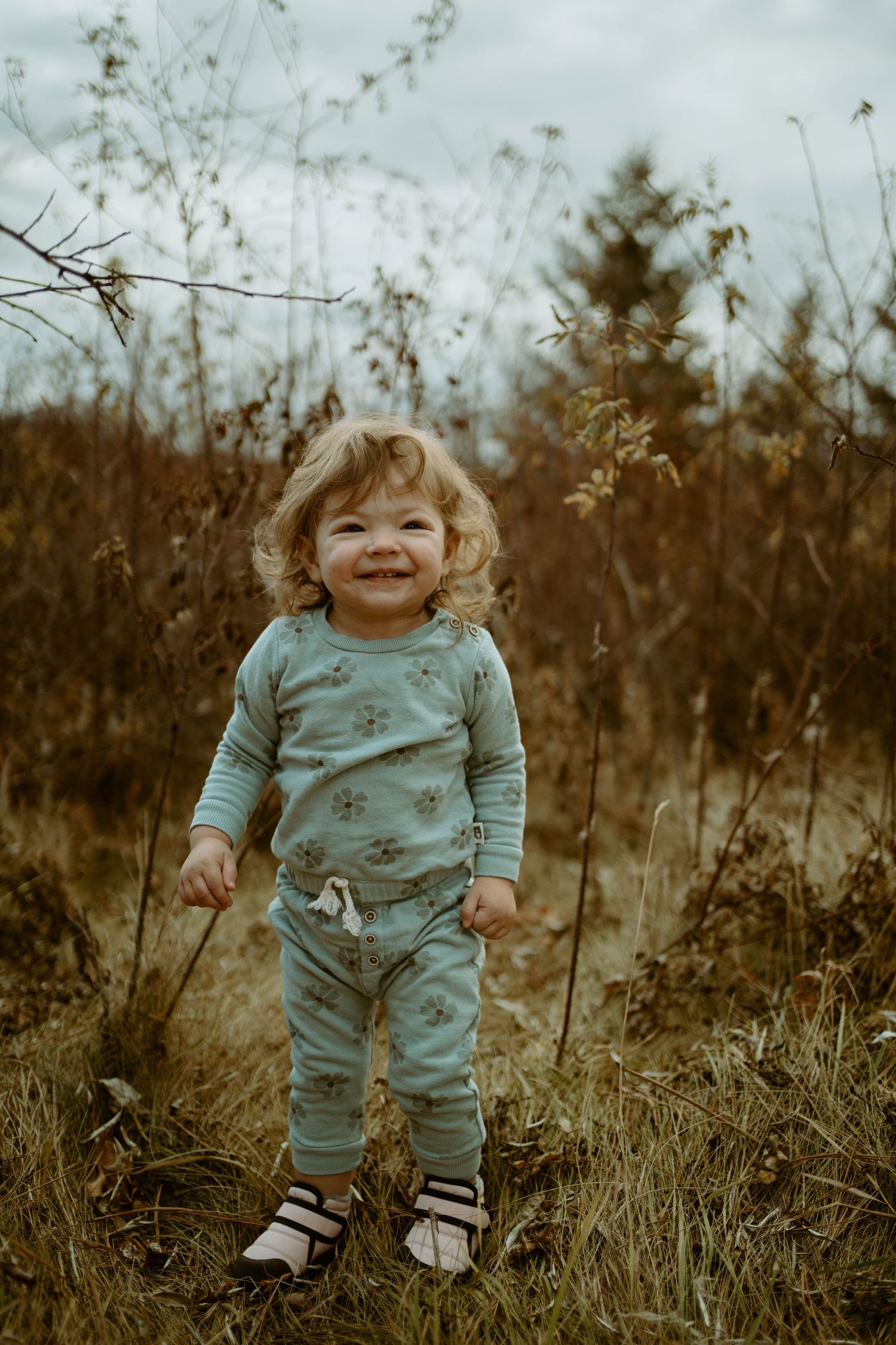 A toddler in blue pajamas smiles while standing in tall grass during golden hour.