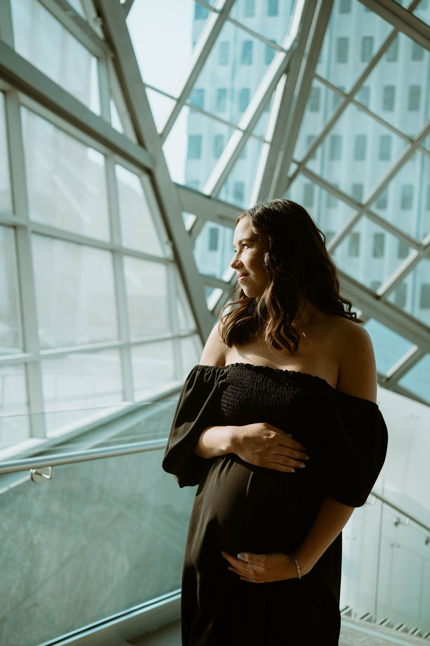 An artistic maternity photo with a woman in black dress posing by modern architecture windows.