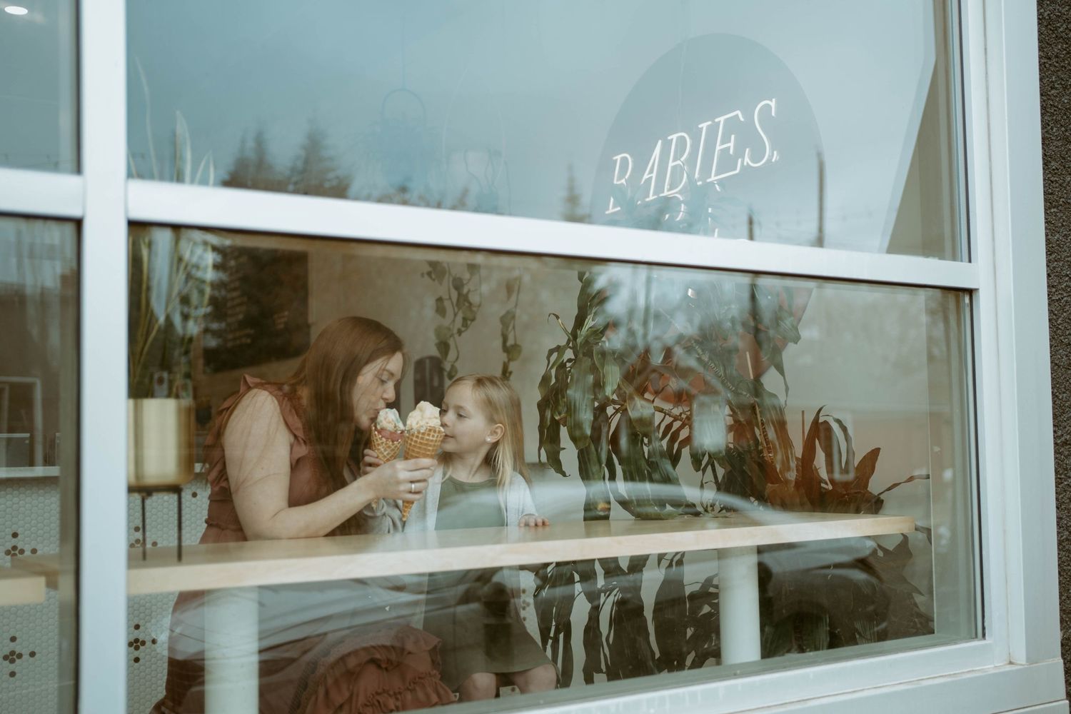 Person enjoying ice cream viewed through window with 'BABIES' sign reflection.