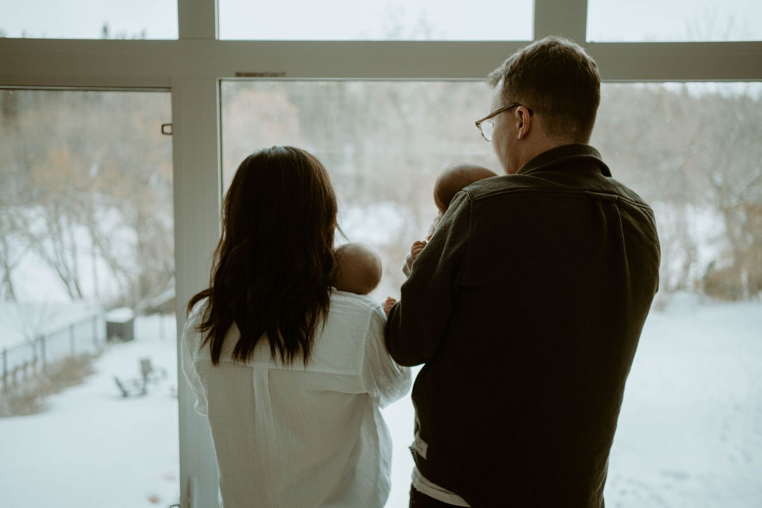 A couple stands at a window with their baby, looking out at a snowy winter landscape.