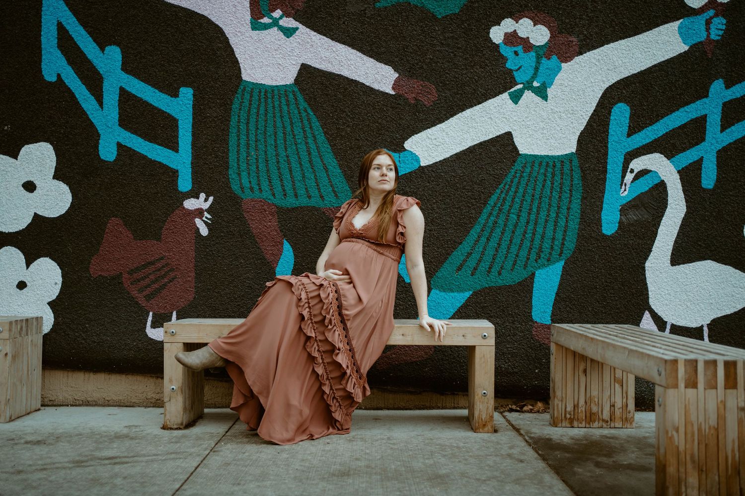 Woman in flowing maxi dress sits on wooden bench against vibrant mural with blue and white designs.