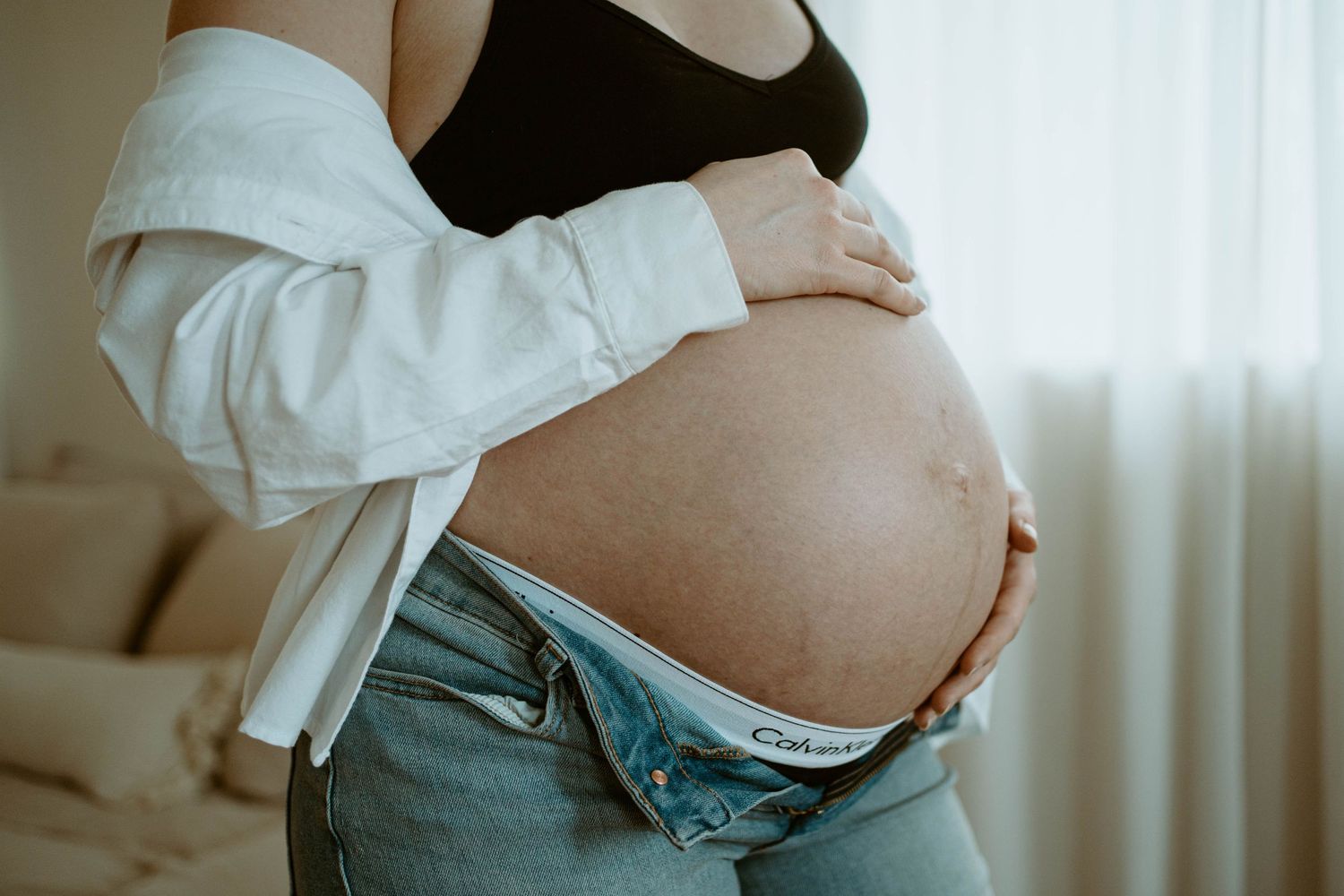 Close-up of a pregnant belly in jeans and white sweater against a window with soft lighting.