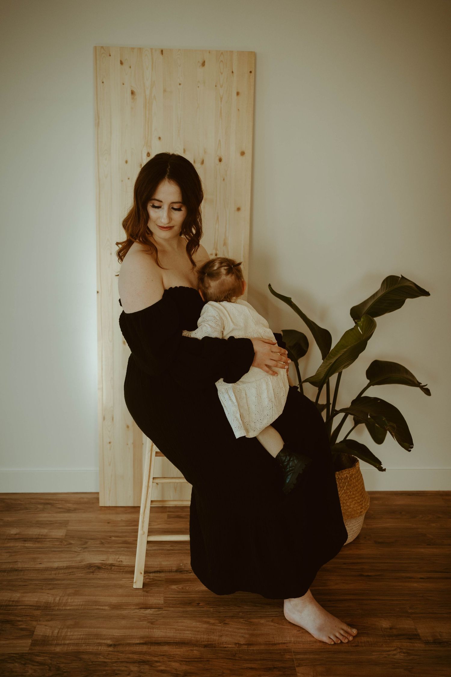 Elegant portrait against wood panel shows tender moment with baby and houseplant in background.