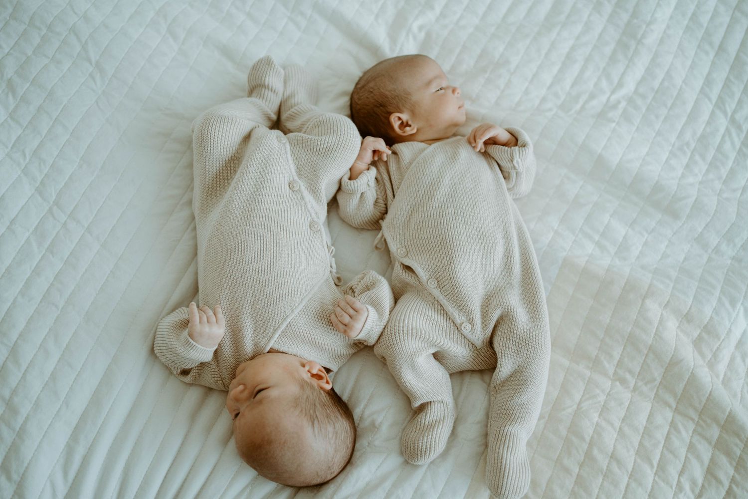 Twin babies in matching cream-colored outfits lying on white quilted bedding.