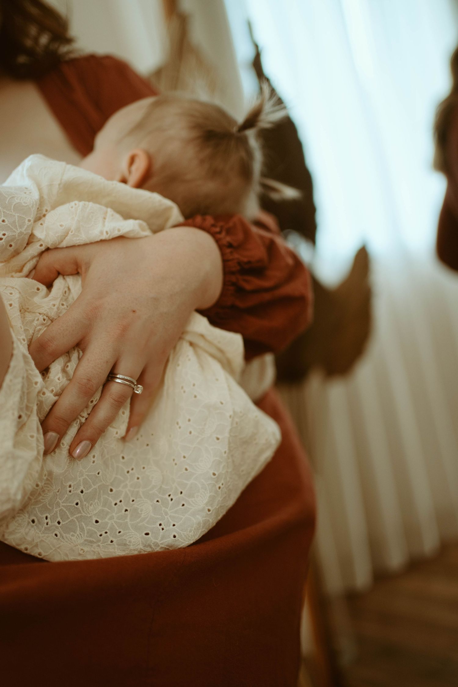 Close-up view of baby in white eyelet dress with caregiver's hand wearing wedding ring visible.