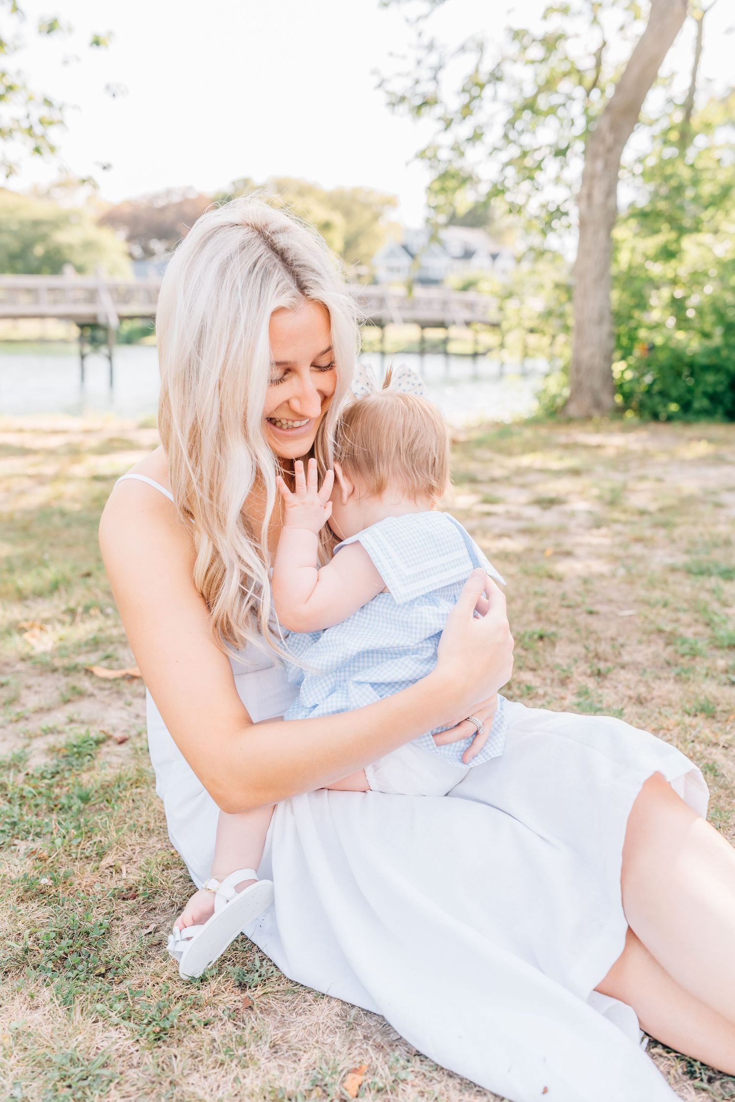 A mother in a pretty white dress embraces a hug from her toddler while sitting in front of a lake at Divine Park.