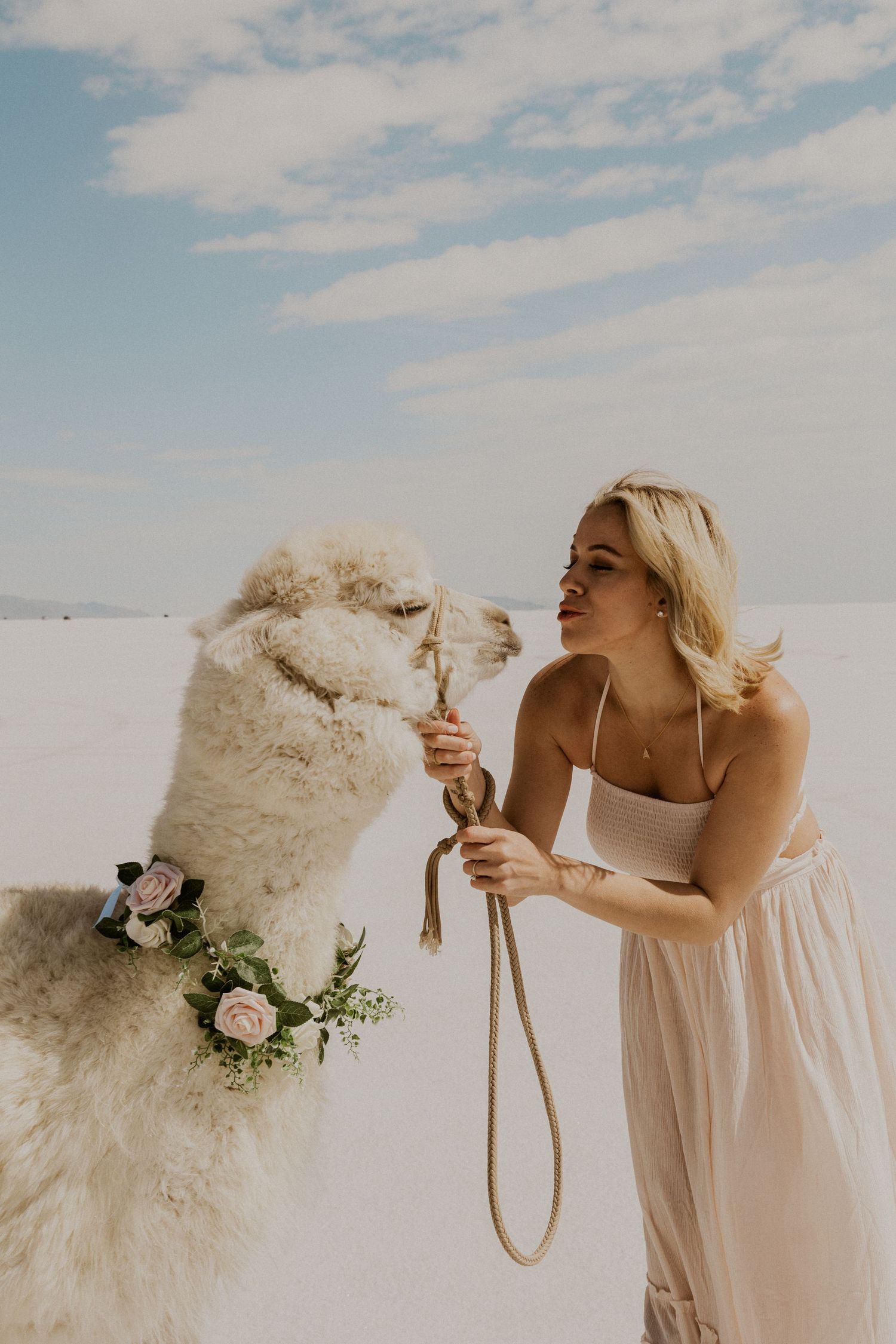 woman in a pink maxi dress holding an alpaca with a floral necklace at the Bonneville Salt Flats, giving a kissy face