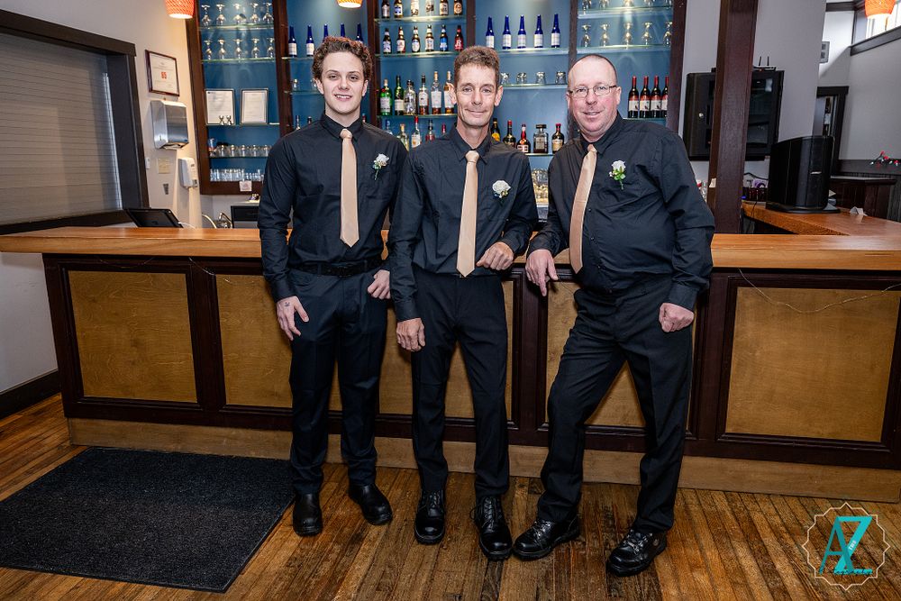 Three groomsmen in black suits pose in front of illuminated bar shelves with wooden accents.