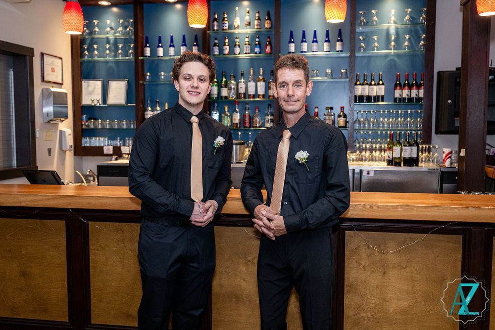 Two groomsmen in black suits pose at bar counter with blue-lit liquor display shelves.