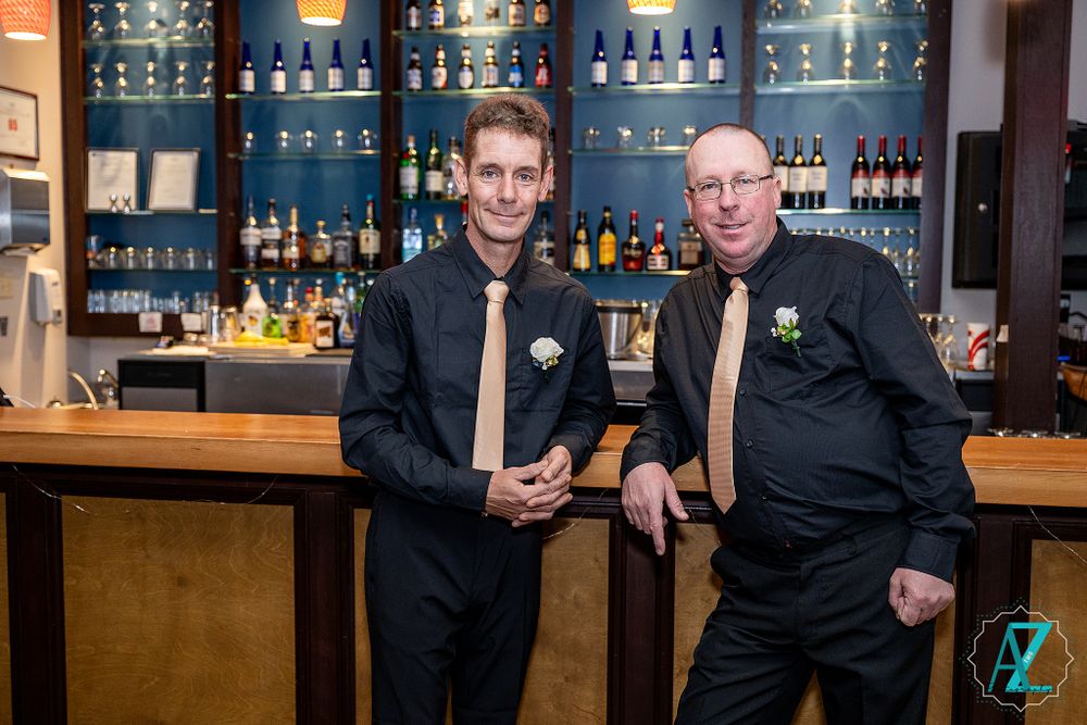 Two members of wedding party in black suits pose at modern bar with blue-lit shelves.