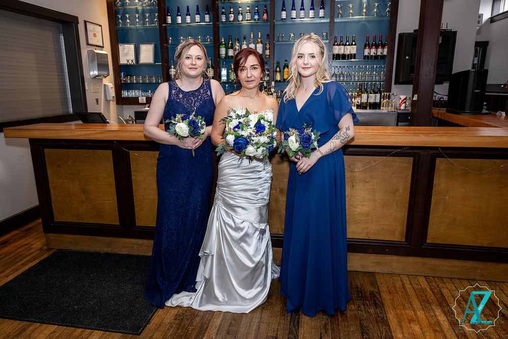 Three women in formal dresses pose at bar counter with blue-lit shelving and wooden accents.