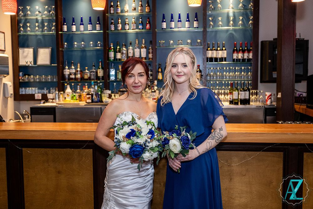 Two women holding floral bouquets pose at elegant bar with illuminated bottle display.