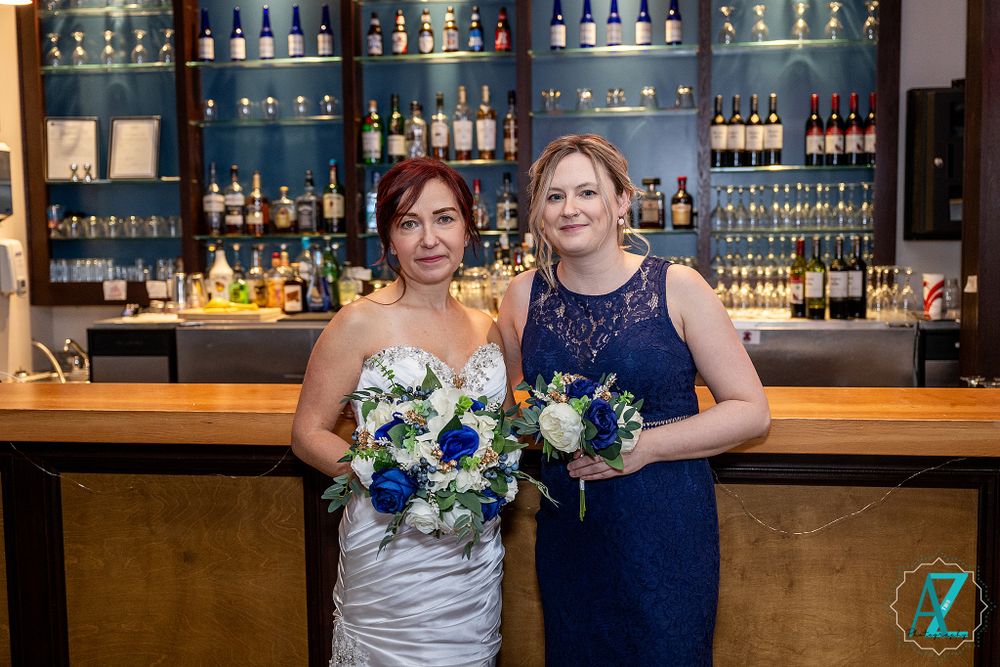 Two women with bouquets pose in formal attire at modern bar with blue-lit bottle displays.
