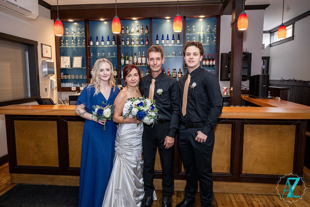 Wedding party members pose in formal attire against blue-lit bar shelving and wooden accents.
