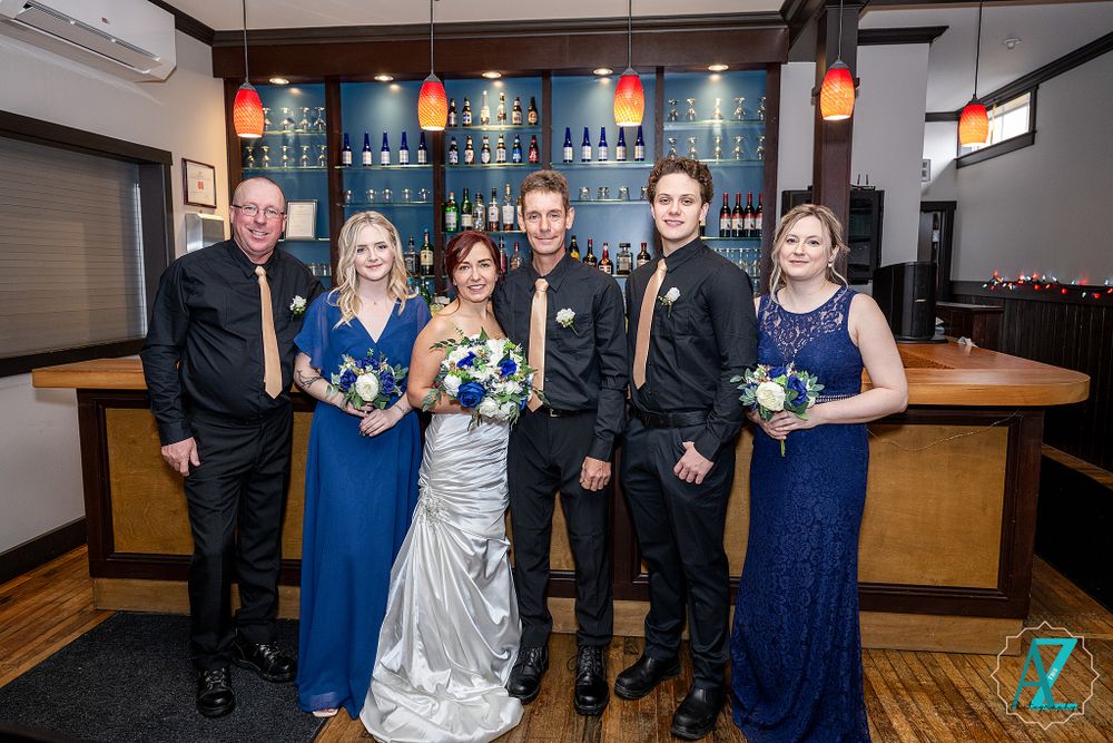 Wedding party poses at bar with blue backdrop and pendant lighting, featuring silver gown and navy blue dresses.