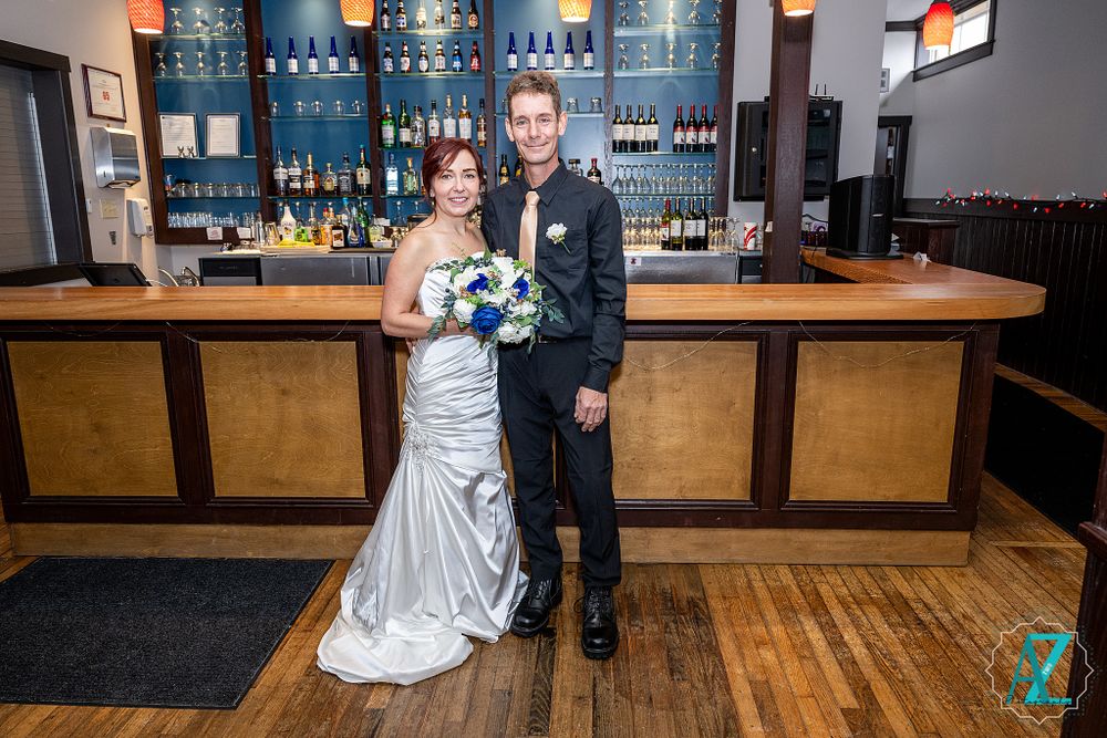 Couple in formal attire stands at wooden bar counter with blue-lit bottle display behind them.