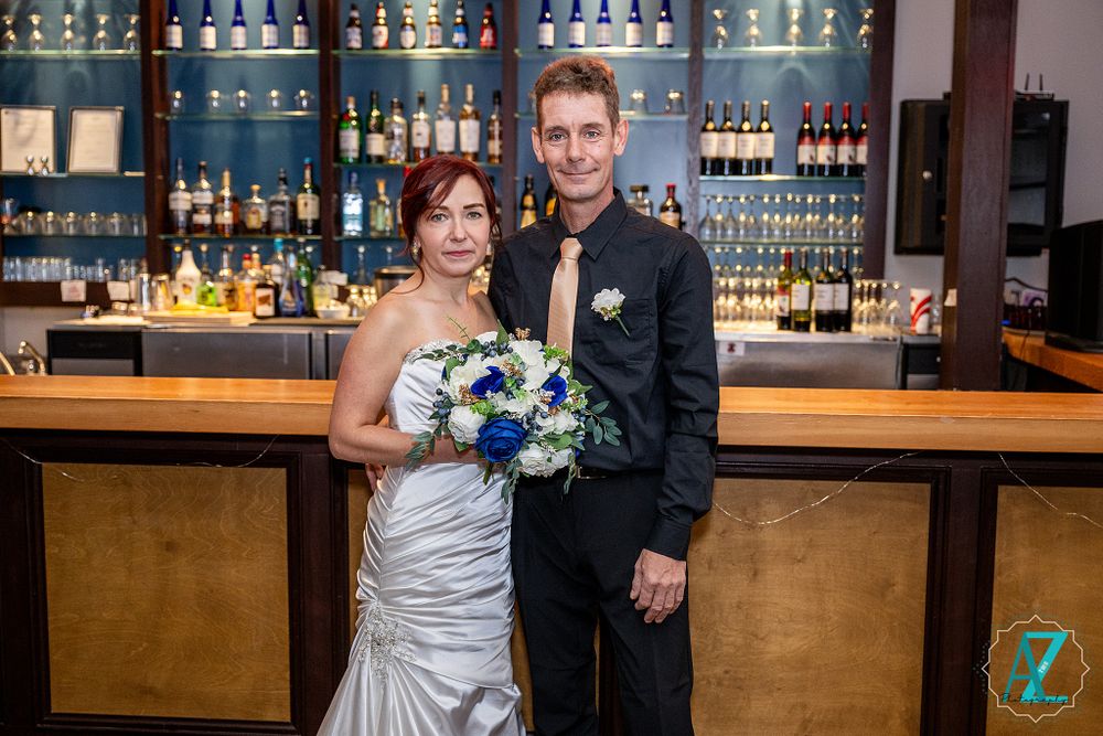 Couple poses at modern bar with illuminated shelving displaying bottles in background.