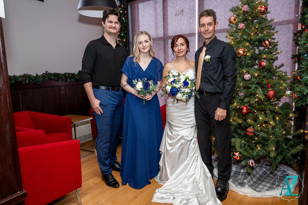 Wedding group in formal attire poses by decorated Christmas tree and red couch.