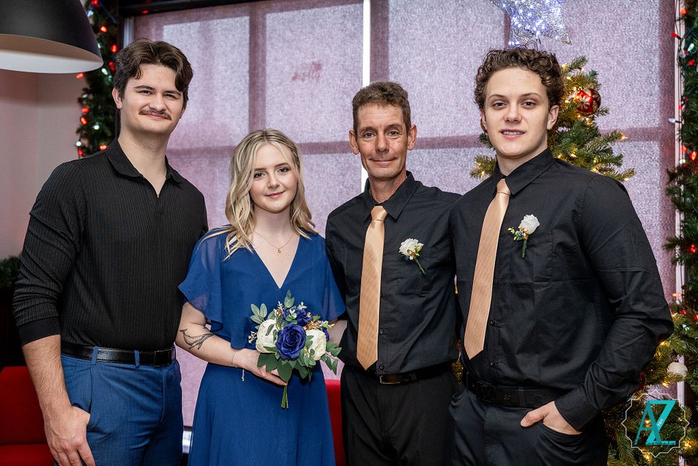 Group celebration photo with Christmas tree and formal attire against pink wall.