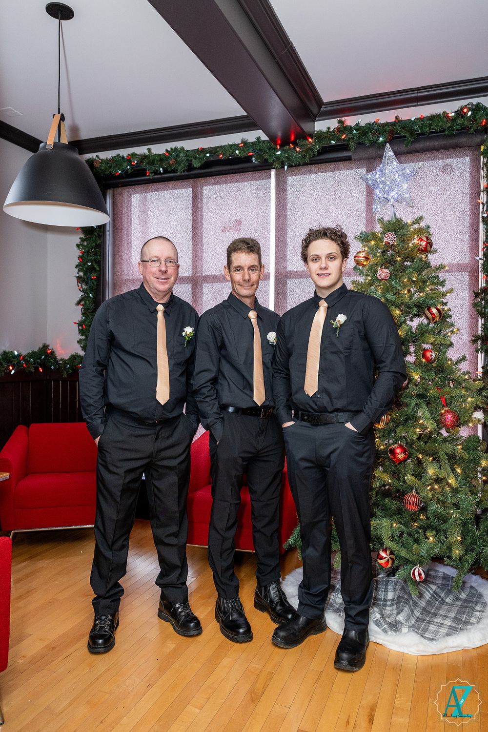 Three formally dressed guests pose near decorated Christmas tree and holiday garland.