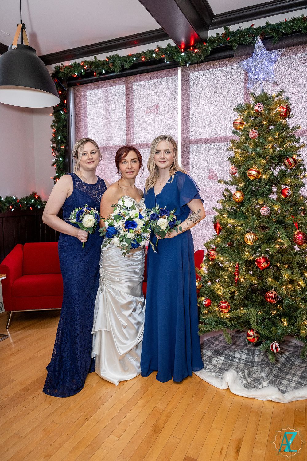 Three women in formal dresses pose with bouquets by Christmas tree.