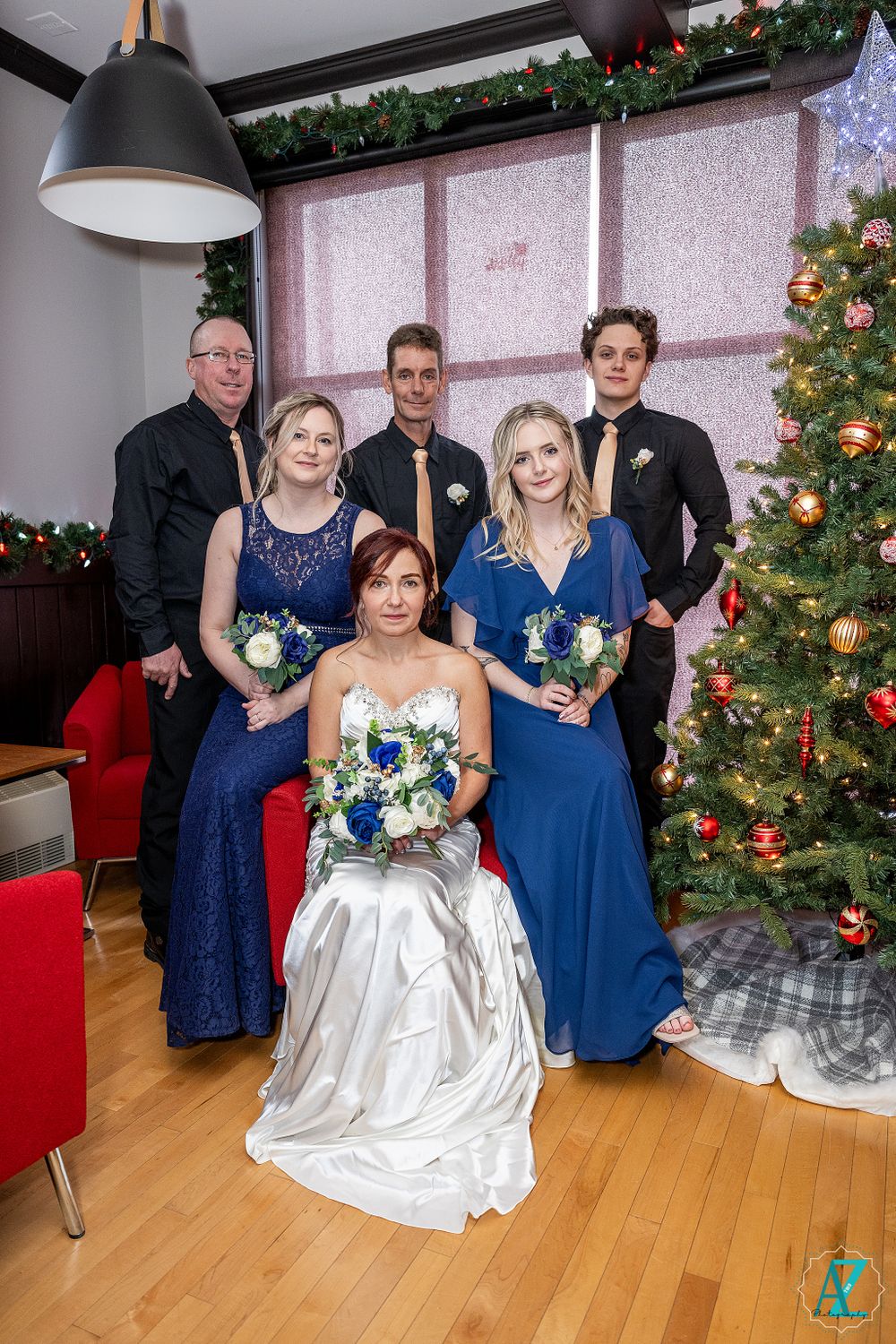 Wedding party poses in front of decorated Christmas tree with bride in white gown and bridesmaids in blue dresses.