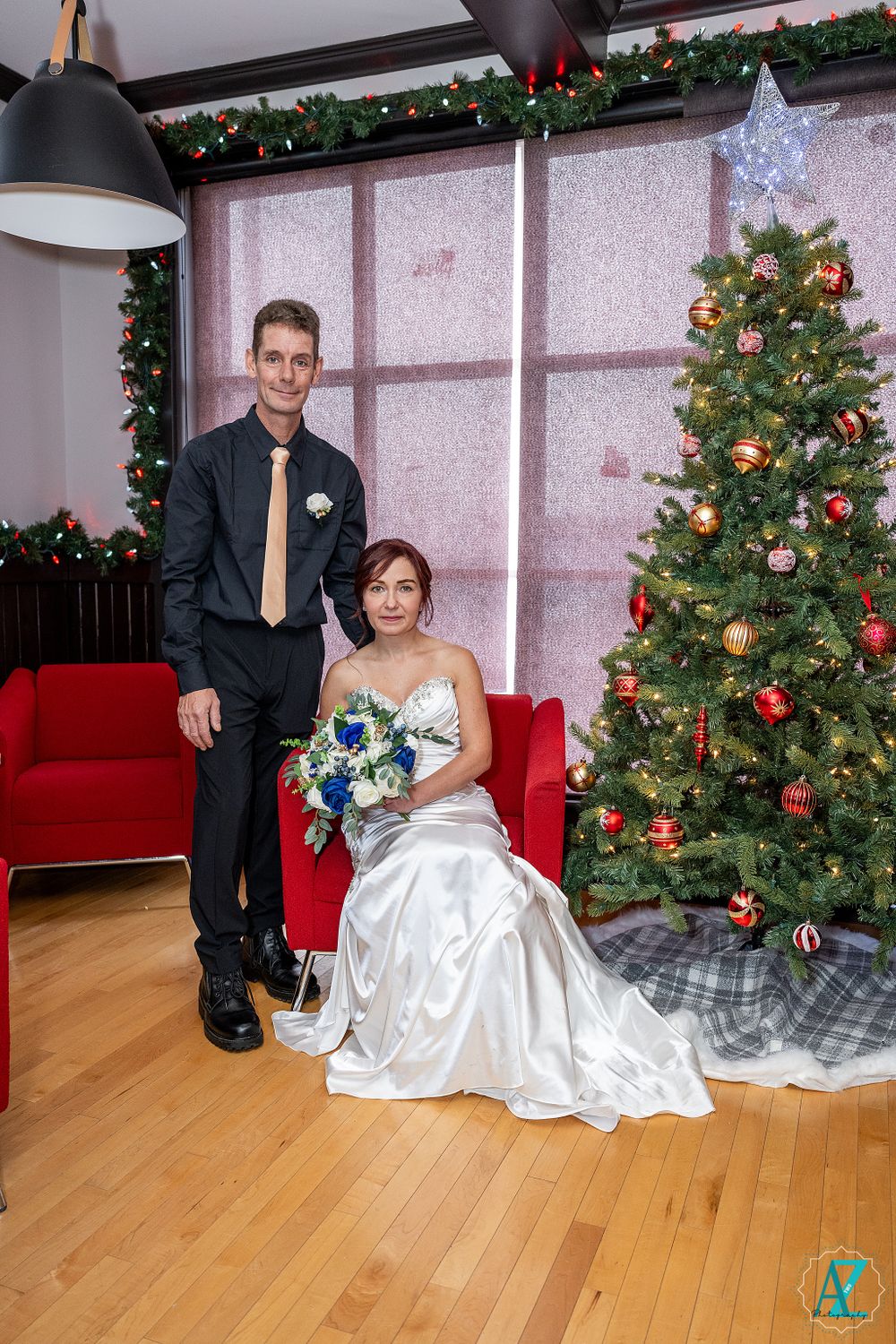 Elegant holiday wedding scene with couple posing by decorated Christmas tree and red couch.