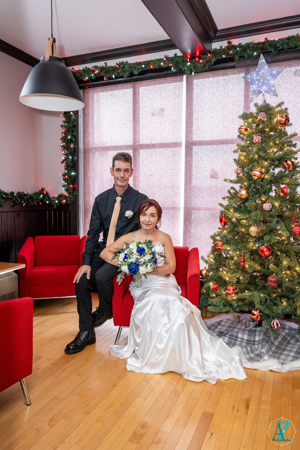 Newlywed couple sits on red couch next to festive Christmas tree in modern setting.