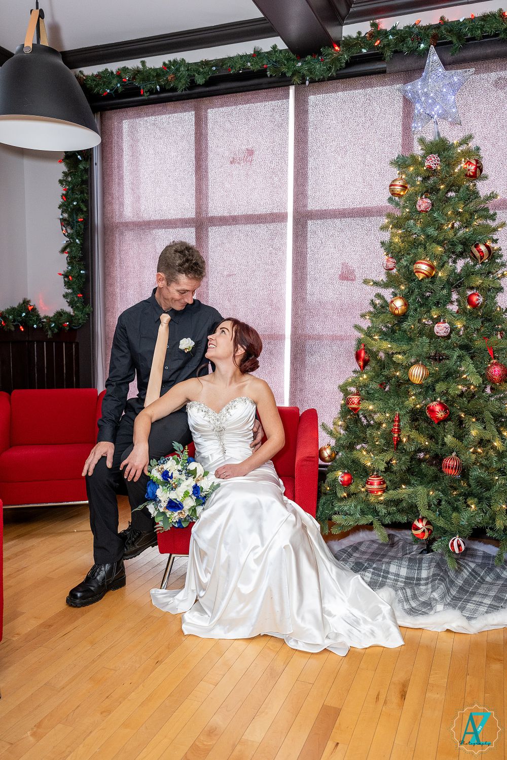 Romantic wedding portrait with couple by Christmas tree and red couch in holiday-decorated room.