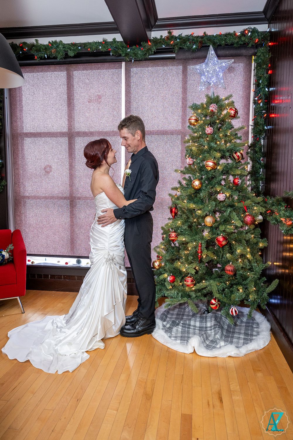Romantic wedding couple embraces in front of decorated Christmas tree in modern indoor setting.