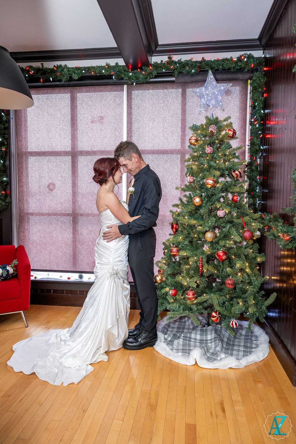 Wedding couple shares intimate moment by decorated Christmas tree in holiday-themed setting.