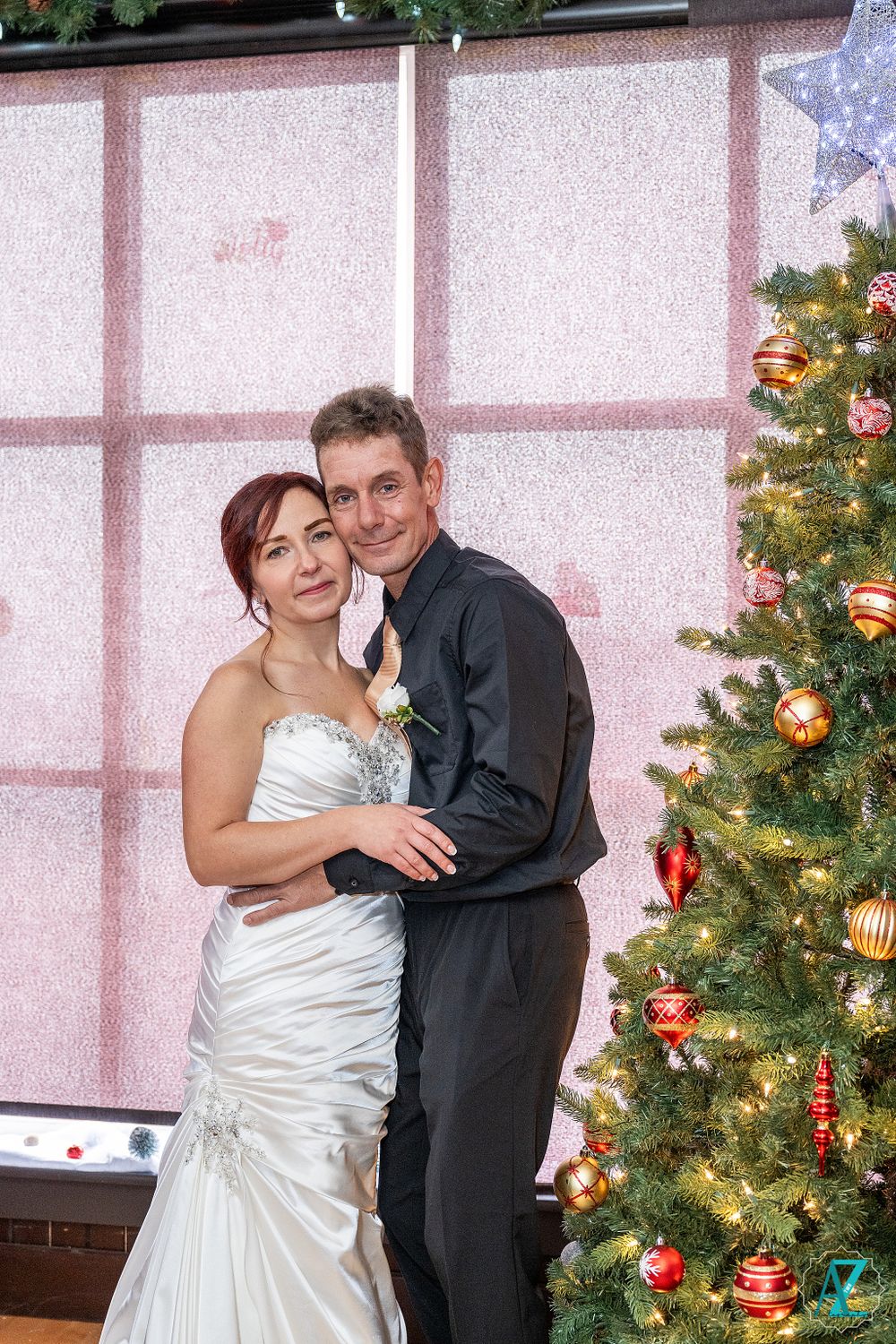 Romantic Christmas wedding portrait with couple by decorated tree and pink backdrop.
