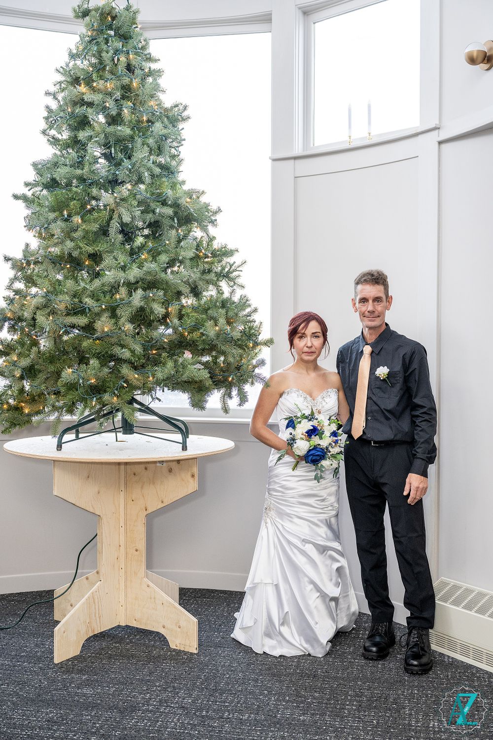 Wedding couple poses next to decorated Christmas tree in white room.
