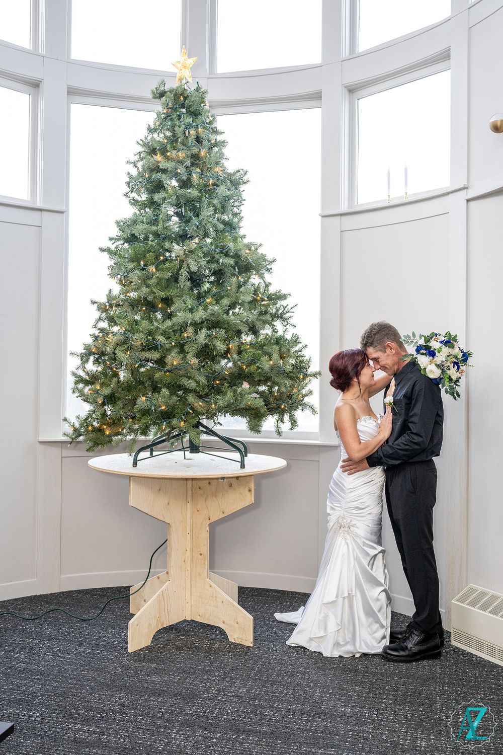 Couple shares intimate moment beside large Christmas tree in curved white room.