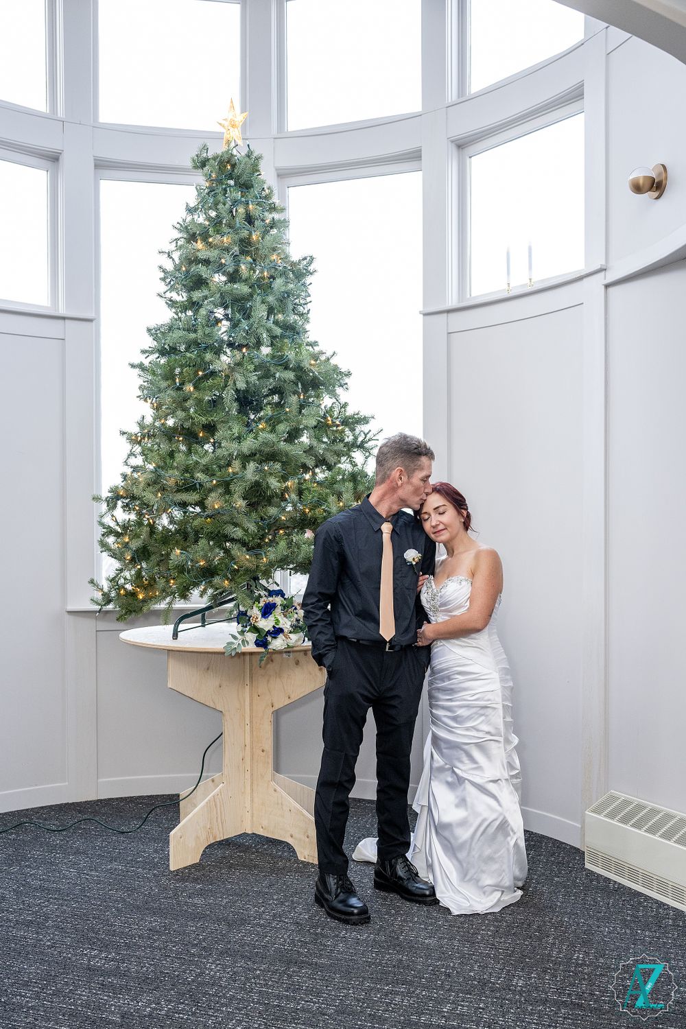 Elegant couple poses beside decorated Christmas tree in curved white room.