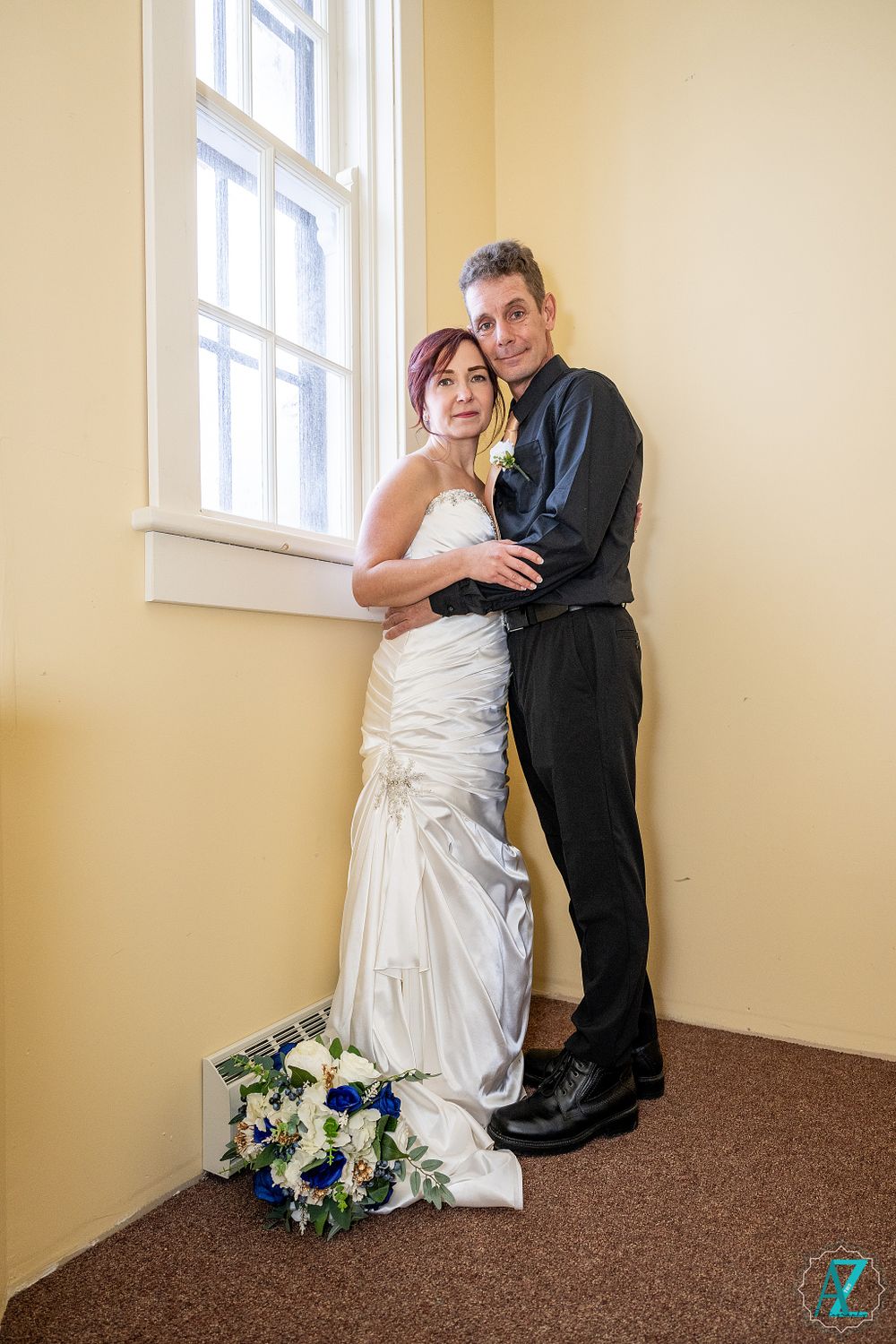 Wedding couple embraces by bright window with floral bouquet.