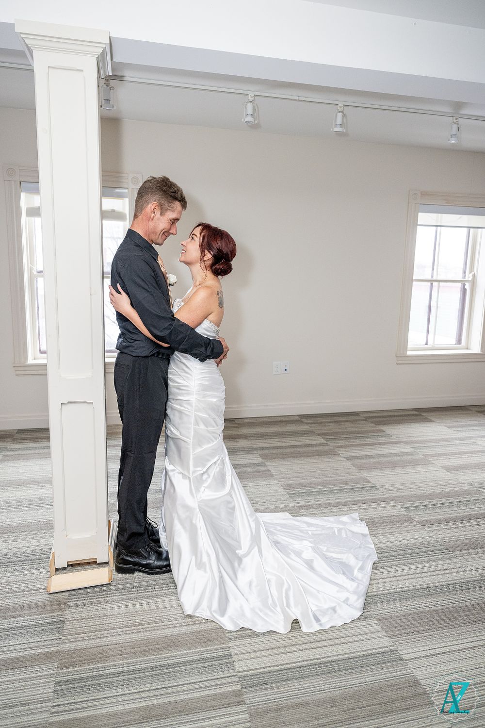 Couple in formal wedding attire embraces near a white column in elegant venue.