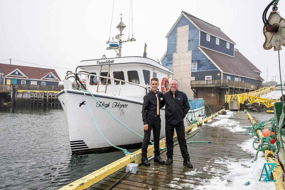 Three people in formal wear pose on dock with fishing boat in harbor.