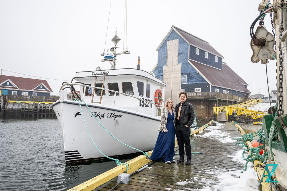Couple in formal attire poses on dock with white fishing boat and blue building in background.