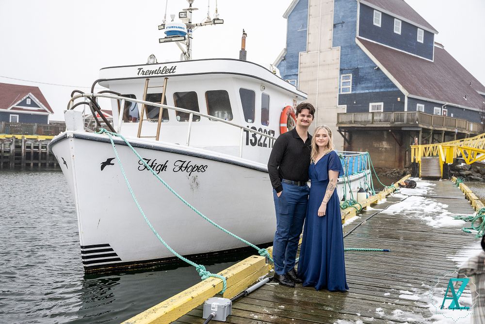 Couple poses on waterfront dock with large white fishing vessel in background.