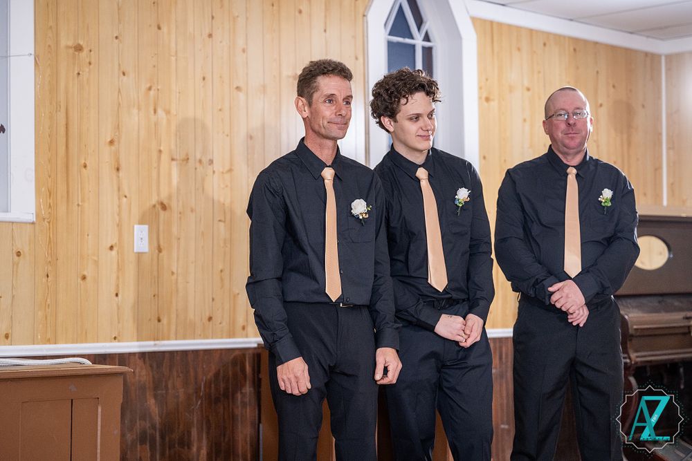 Three groomsmen in black suits with gold ties stand together during a wedding ceremony in a wood-paneled room.