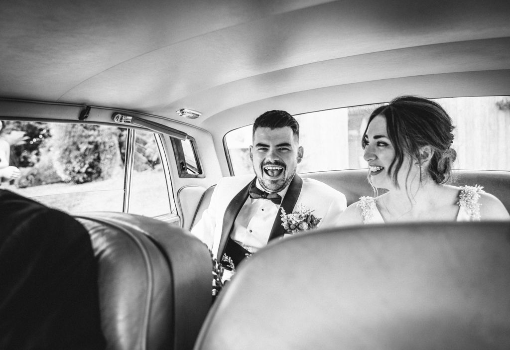 Black and white photo of newlyweds laughing together in classic car backseat.