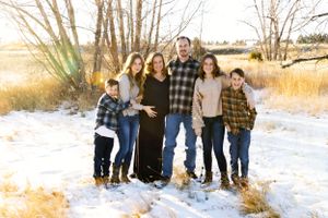 A family stands together on snowy ground at sunset, with bare winter trees and golden grasses in the background.